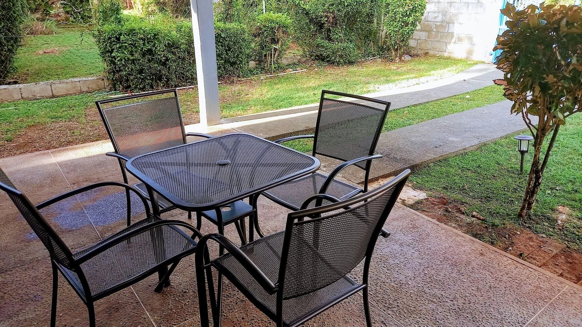 Un patio al aire libre con una mesa y sillas de metal, rodeado de un jardín y un sendero.