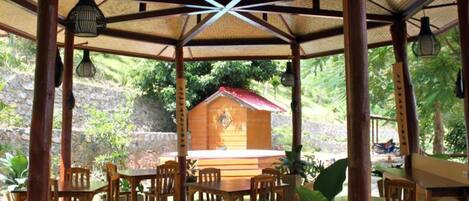 A covered outdoor seating area with wooden tables and chairs, a central wooden structure, and hanging light fixtures.
