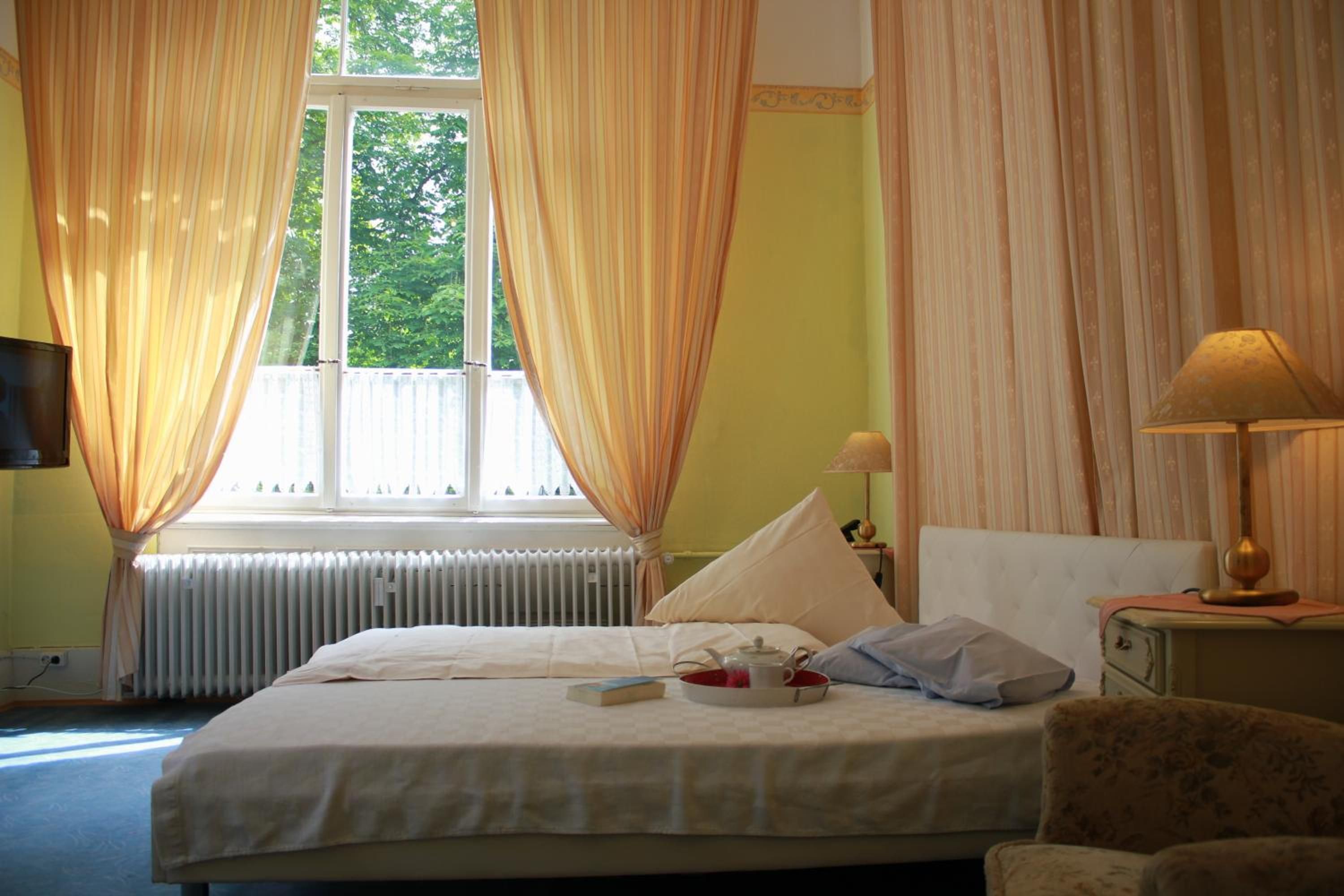 A hotel room with a bed, a nightstand with a lamp, a window with curtains, and a radiator.
