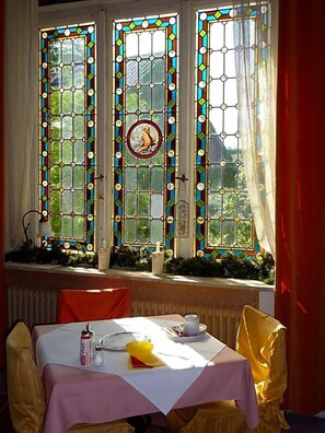 A dining area with a table set for two, a window with stained glass, and a view of greenery outside.