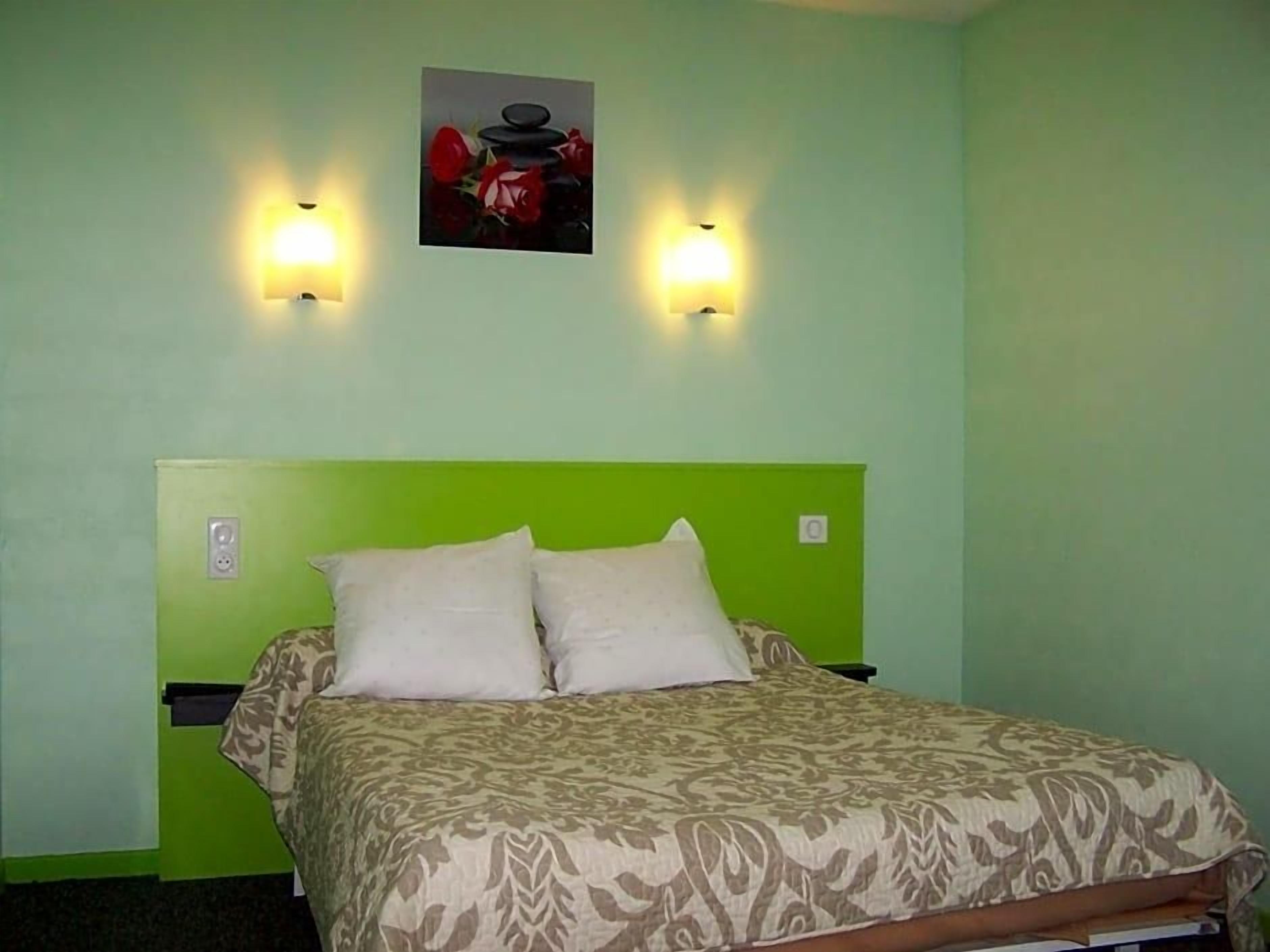 A room with a green wall, a bed with a patterned bedspread, two wall-mounted lights, and a framed picture above the bed.