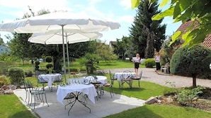 Un patio al aire libre con mesas y sillas, rodeado de follaje y un sendero.