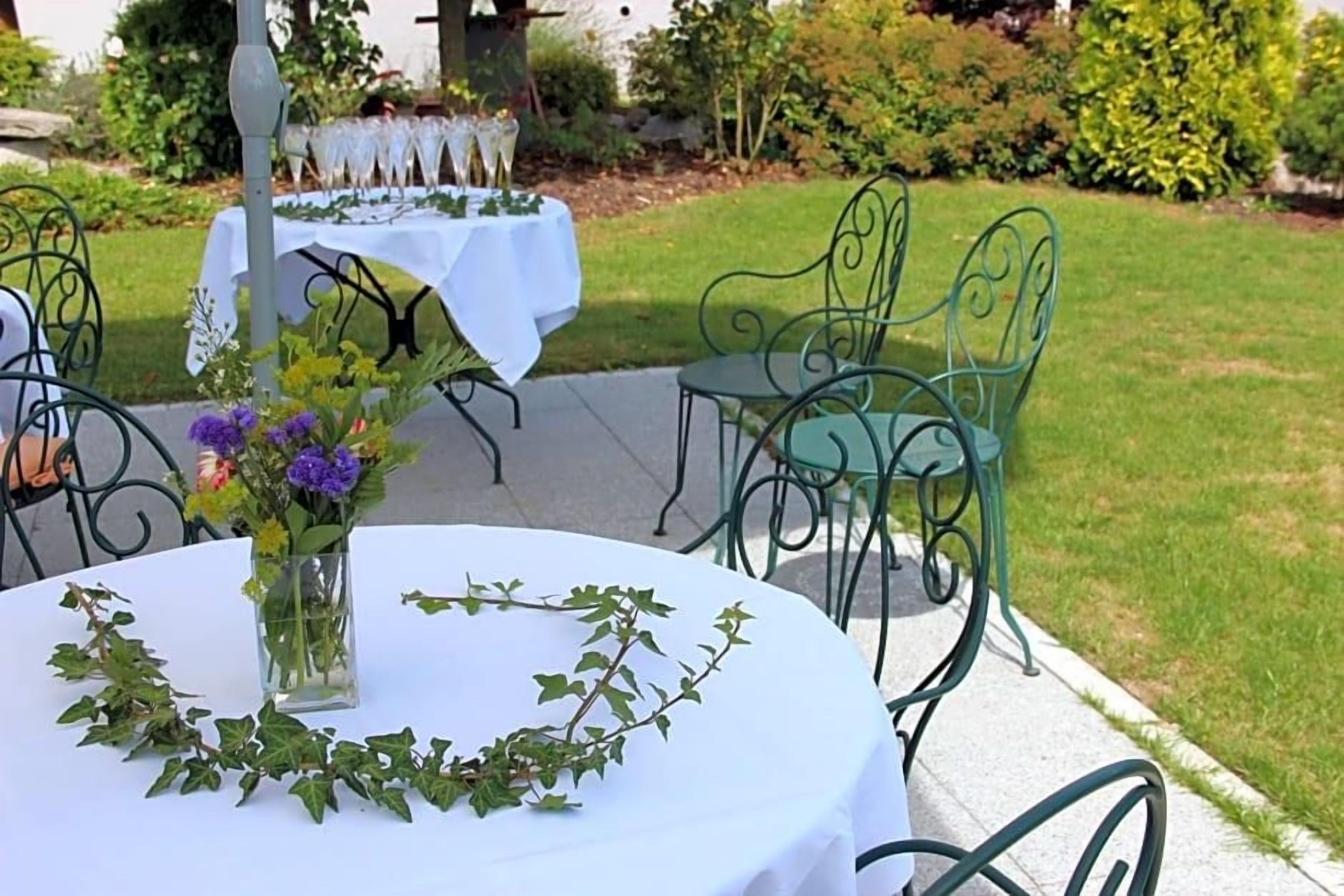 A round table with a white tablecloth, surrounded by green metal chairs, set outdoors with a garden in the background.