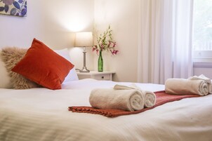Una cama bien hecha con toallas enrolladas, una almohada roja, un jarrón con flores y una lámpara en una mesita de noche.