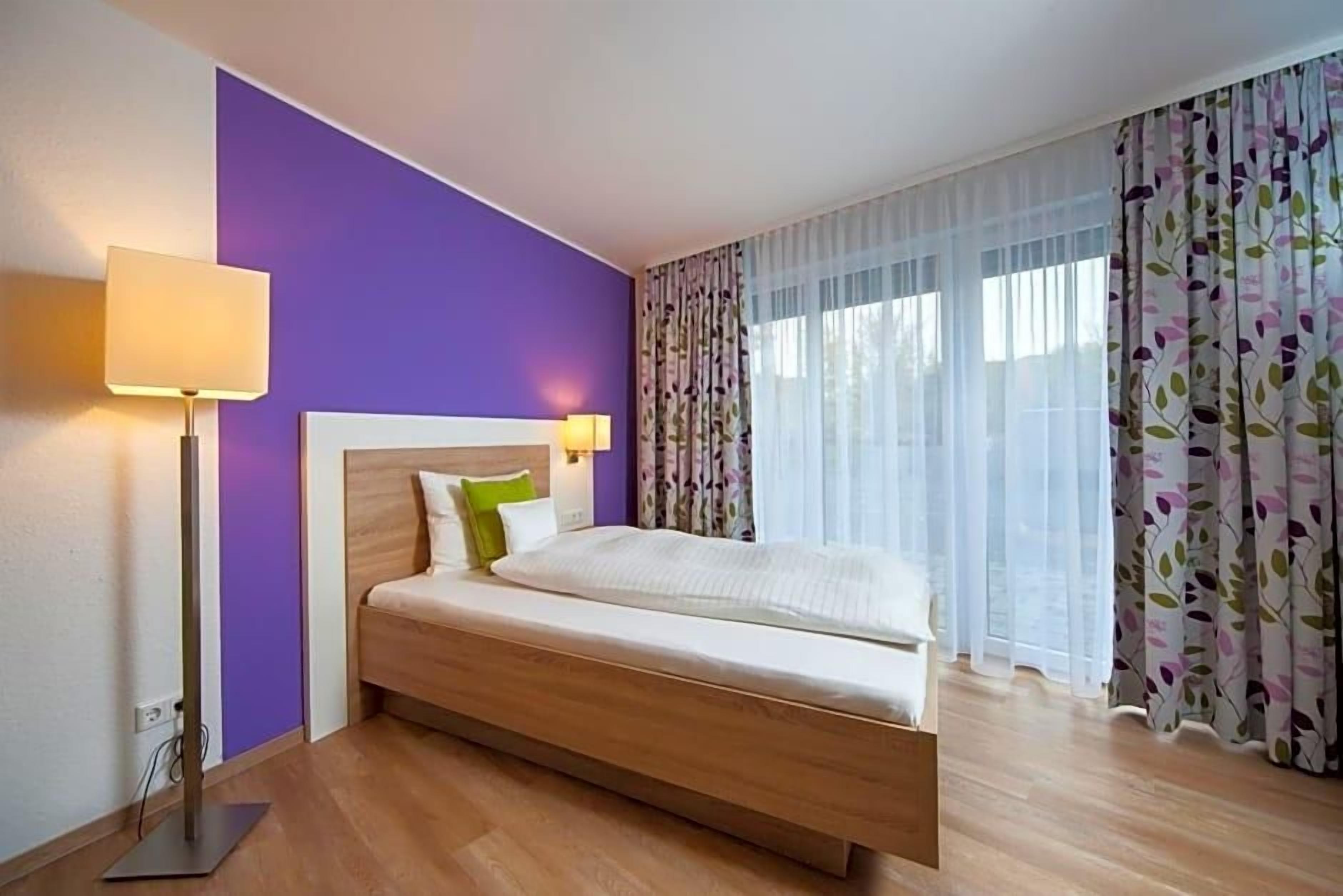 A hotel room with a bed, a nightstand, a lamp, and a window with curtains.