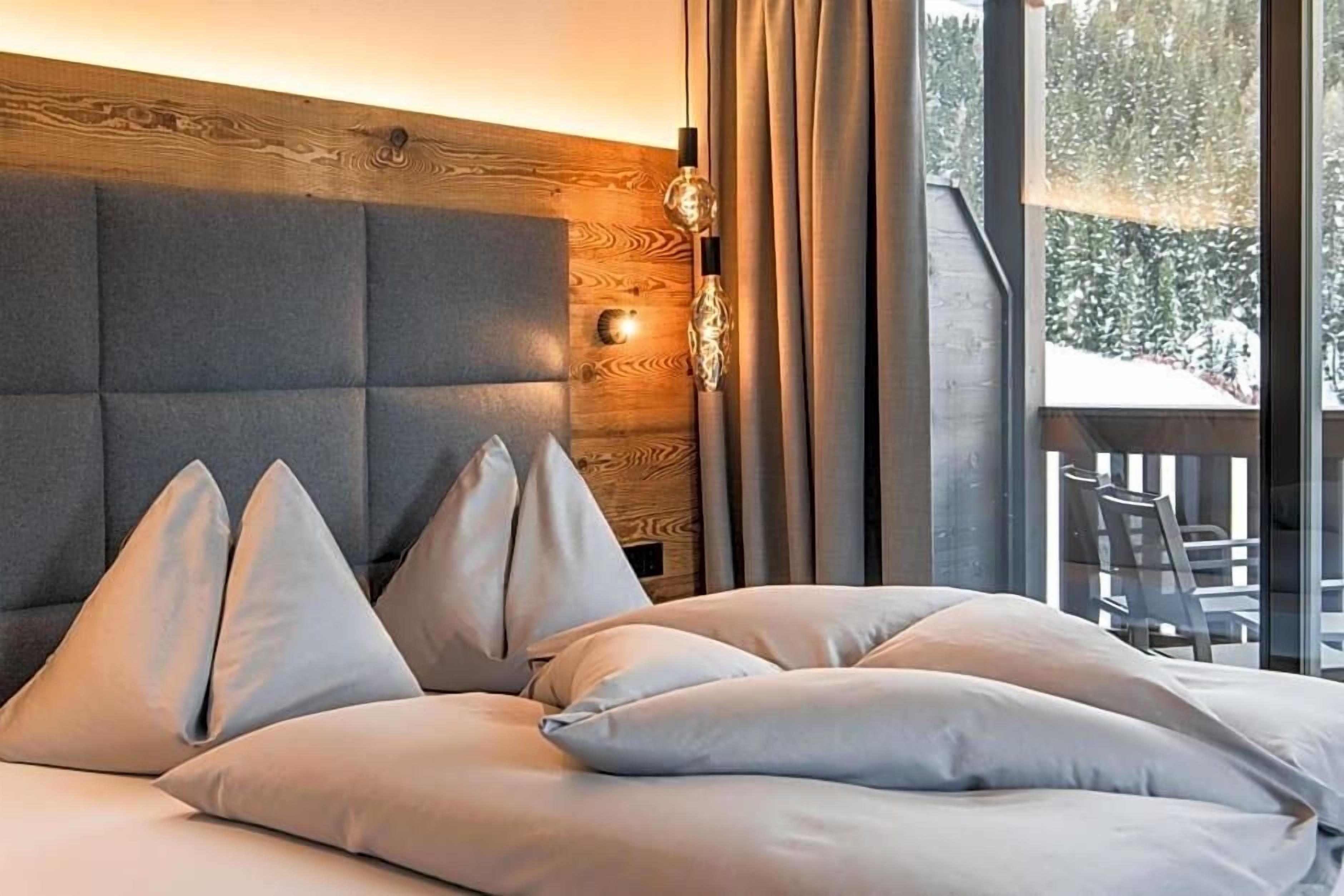 Un dormitorio con cabecera de madera, cama con múltiples almohadas, mesita de noche con lámpara y vista a un paisaje nevado a través de una ventana.