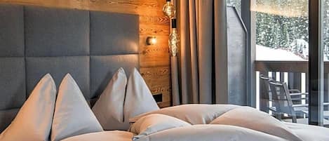 Un dormitorio con cabecera de madera, cama con múltiples almohadas, mesita de noche con lámpara y vista a un paisaje nevado a través de una ventana.