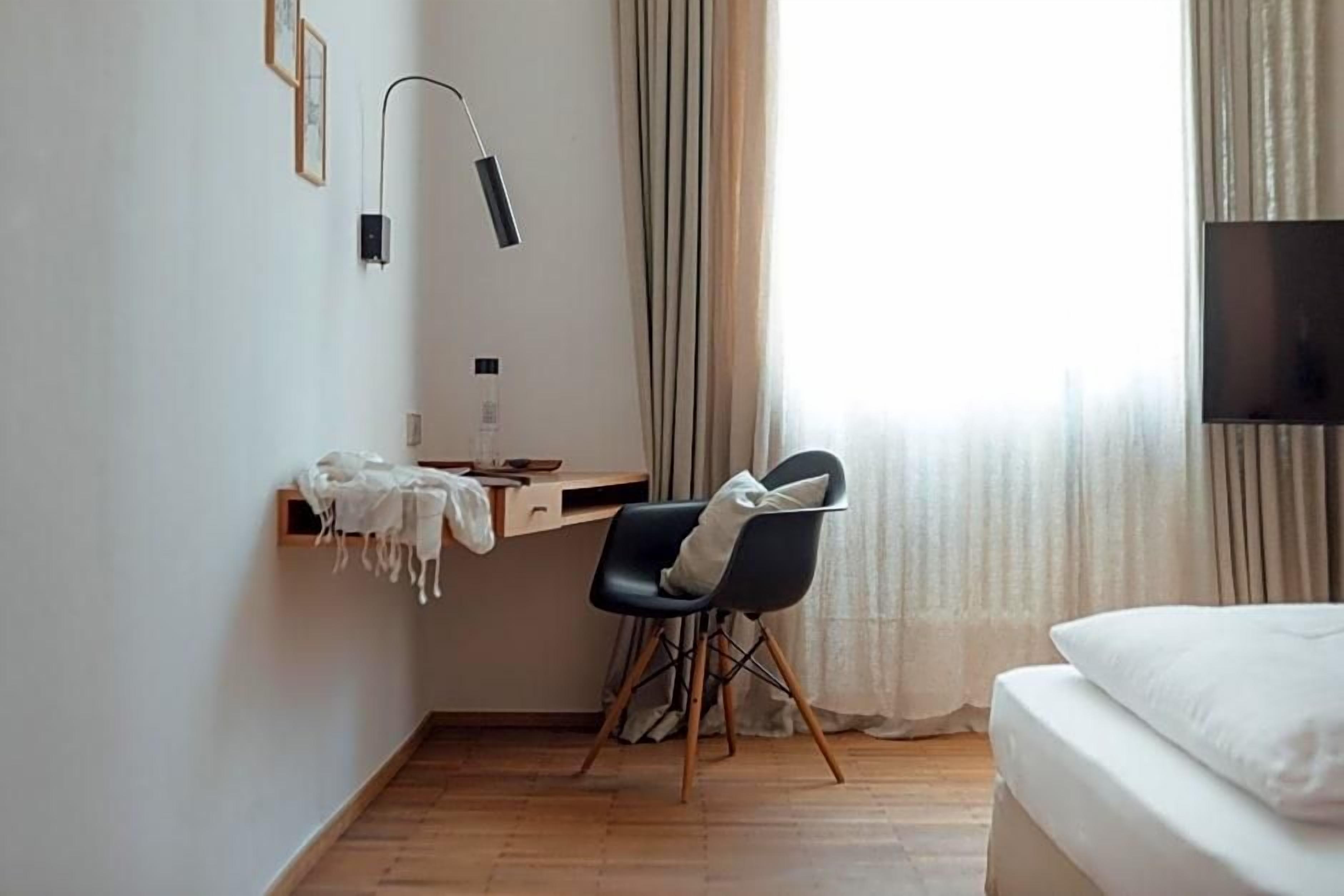 Habitación de hotel con cama, escritorio, silla, lámpara y ventana con cortinas.