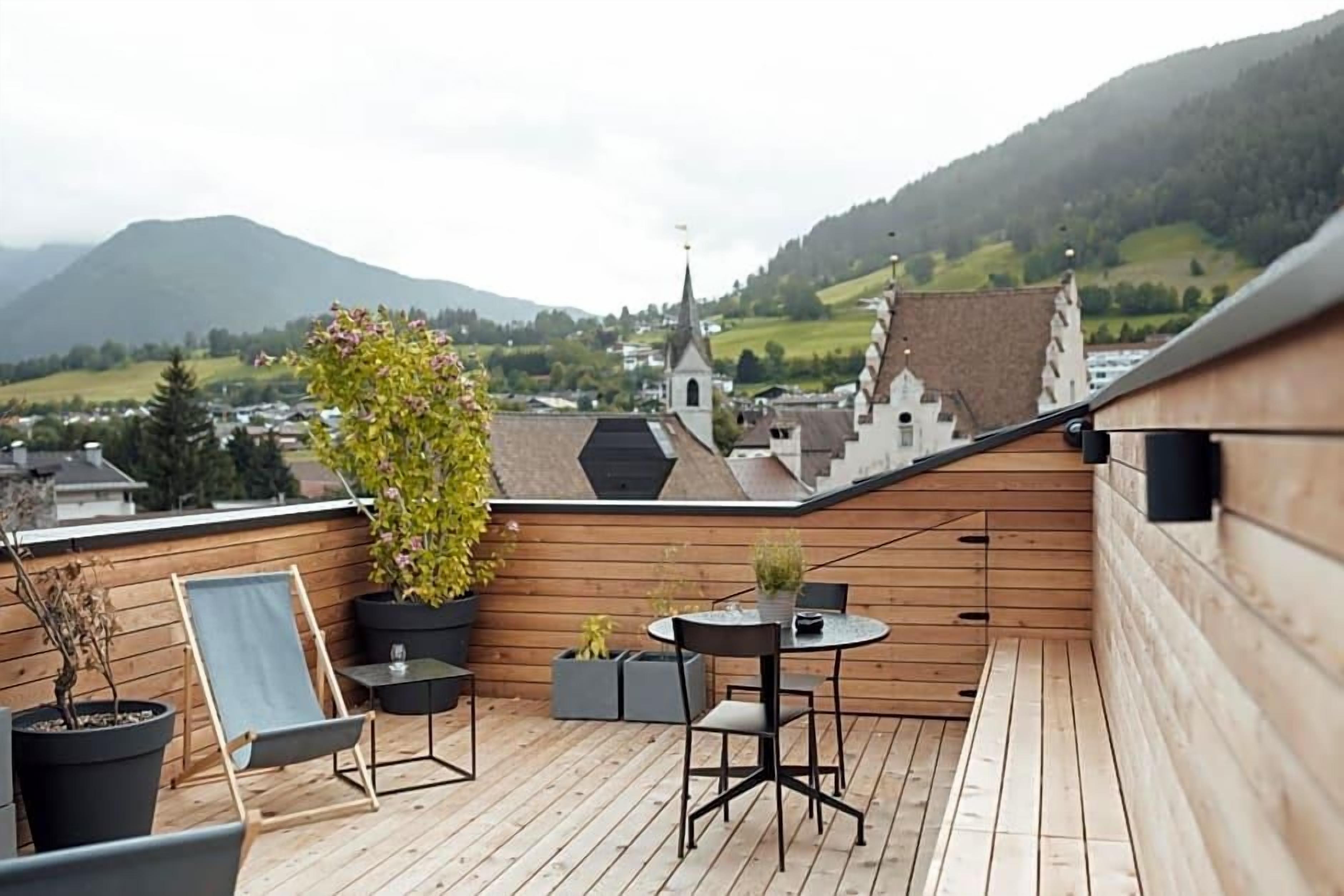 Une terrasse en bois avec une table et des chaises, des plantes en pot, et une vue sur une ville et des montagnes en arrière-plan.