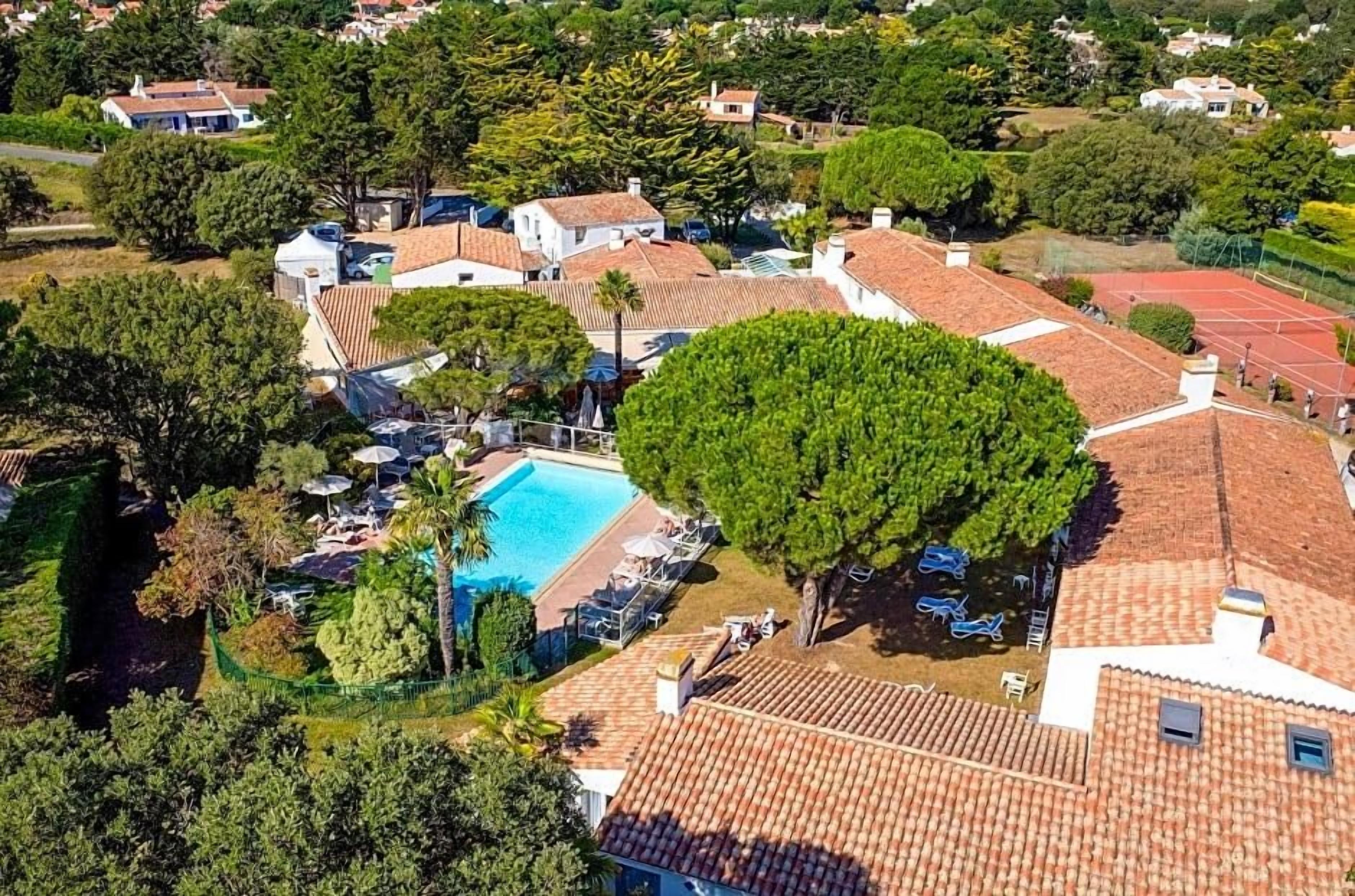 A residential area with a swimming pool surrounded by houses with red-tiled roofs, trees, and a tennis court.