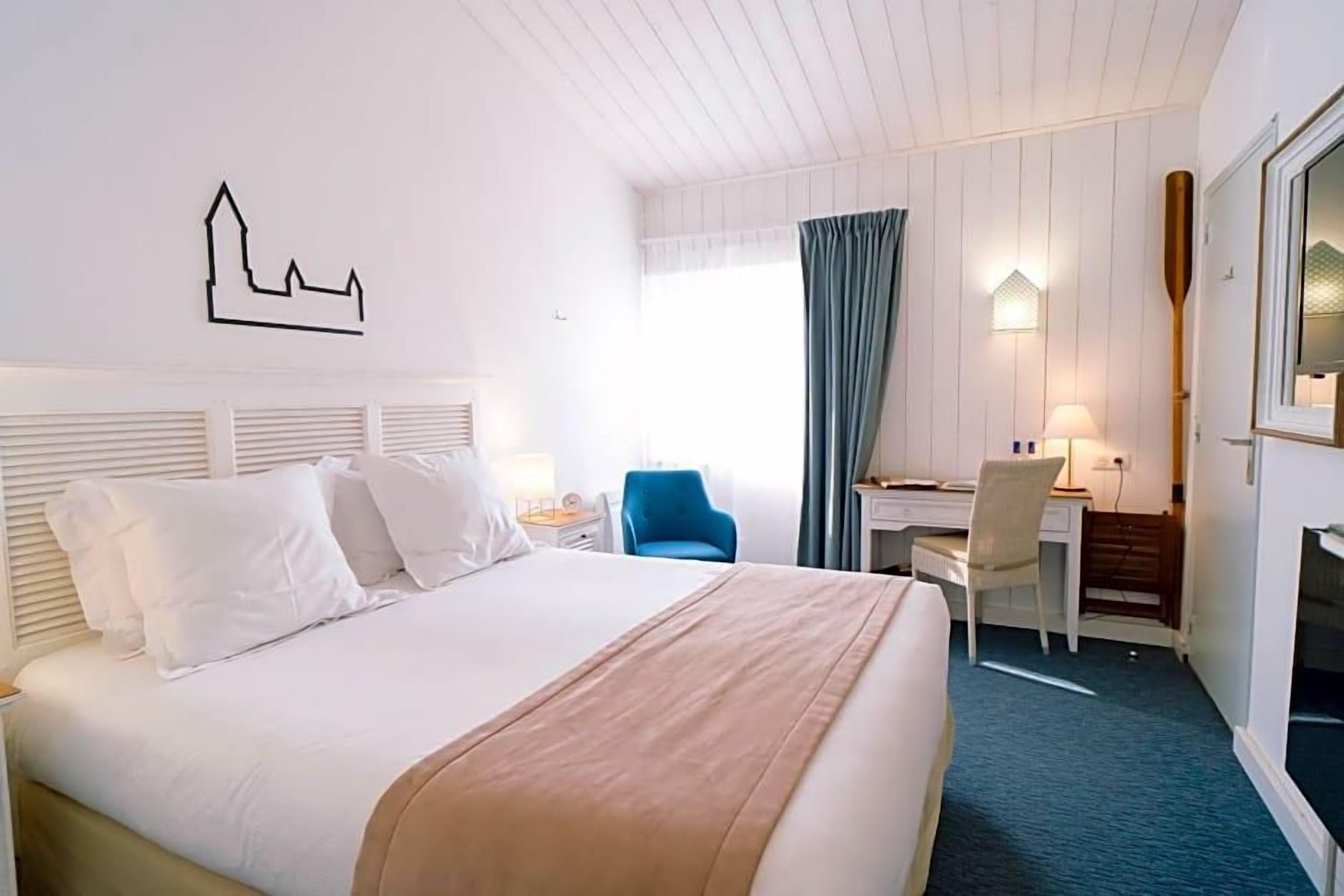 A hotel room with a bed, a desk, a chair, a lamp, and a wall-mounted artwork.