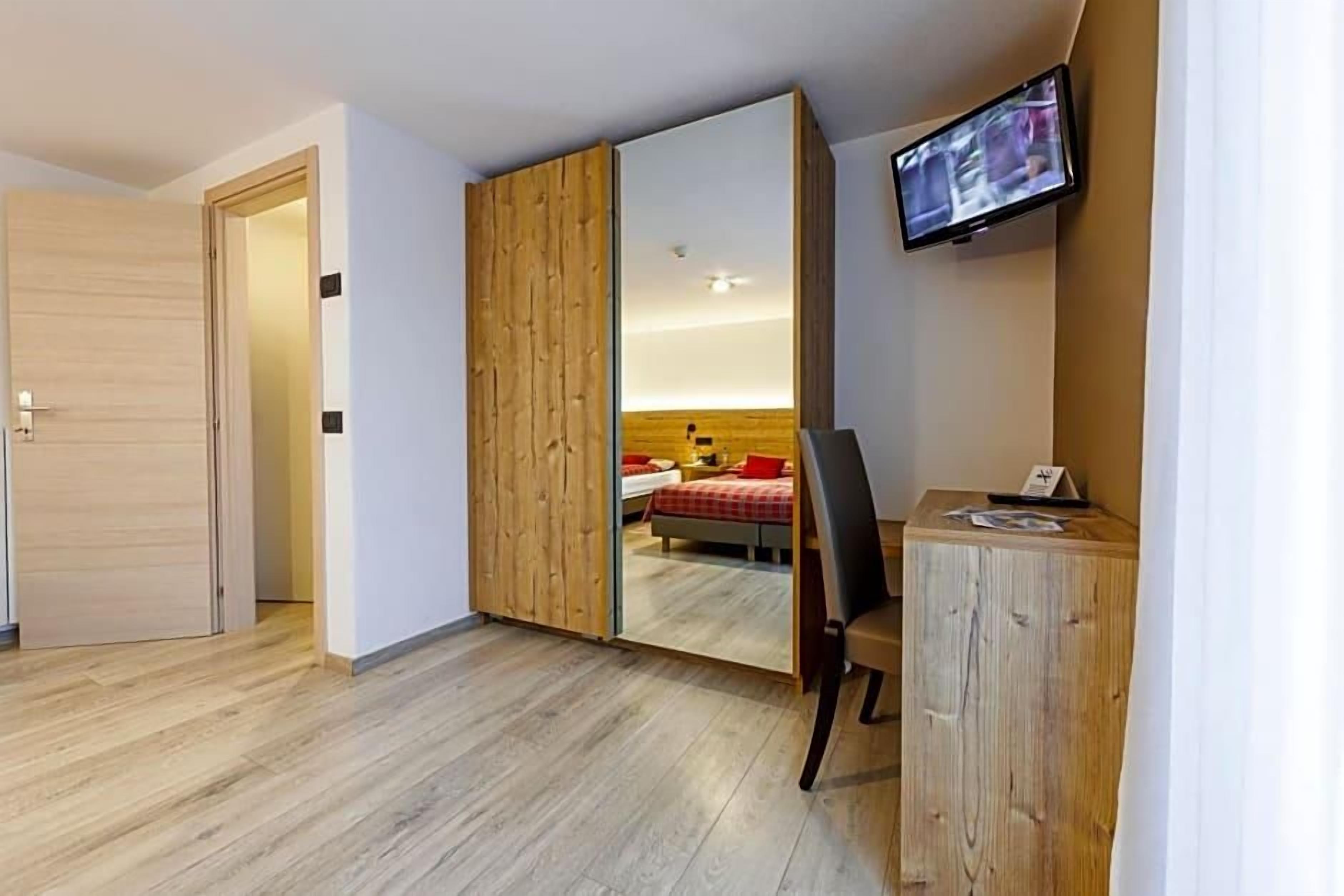 Une chambre d’hôtel avec une armoire en bois, un bureau avec une chaise, un téléviseur fixé au mur et un lit avec une literie rouge.