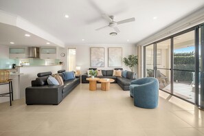 A modern living room with a sofa, coffee table, and chairs, a kitchen area with a fridge and stove, and a balcony with a view. - Waters Edge Twin Waters - Pool, Deck & Kayaks (Coolum Region)