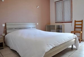 Un dormitorio con cama, mesita de noche, silla, ventana con cortinas y estantería.