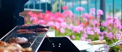 Una parrilla con varios tipos de carne asándose al aire libre, con montañas y flores rosadas al fondo.