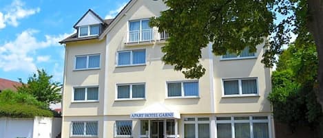 Un hotel de varios pisos con fachada plana, un balcón en el segundo nivel y un cartel que indica que es un 'APART HOTEL GARNI'.