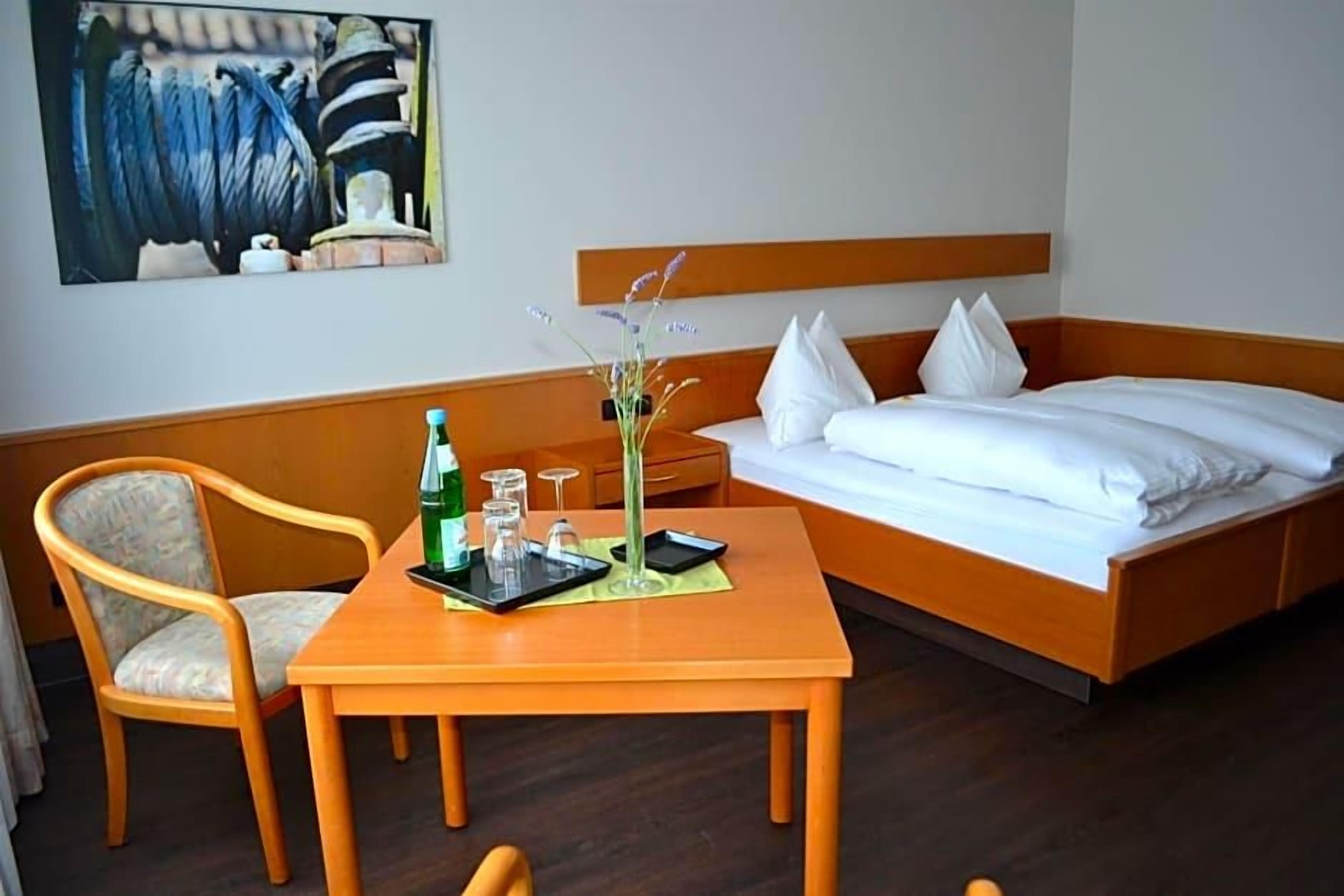 Habitación de hotel con cama, una mesa de madera con una botella, vasos y un florero con flores.