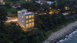 A modern multi-story building with a glass facade, situated on a rocky shoreline with dense greenery. - ?? ???? ???? ?? (Yeosu)