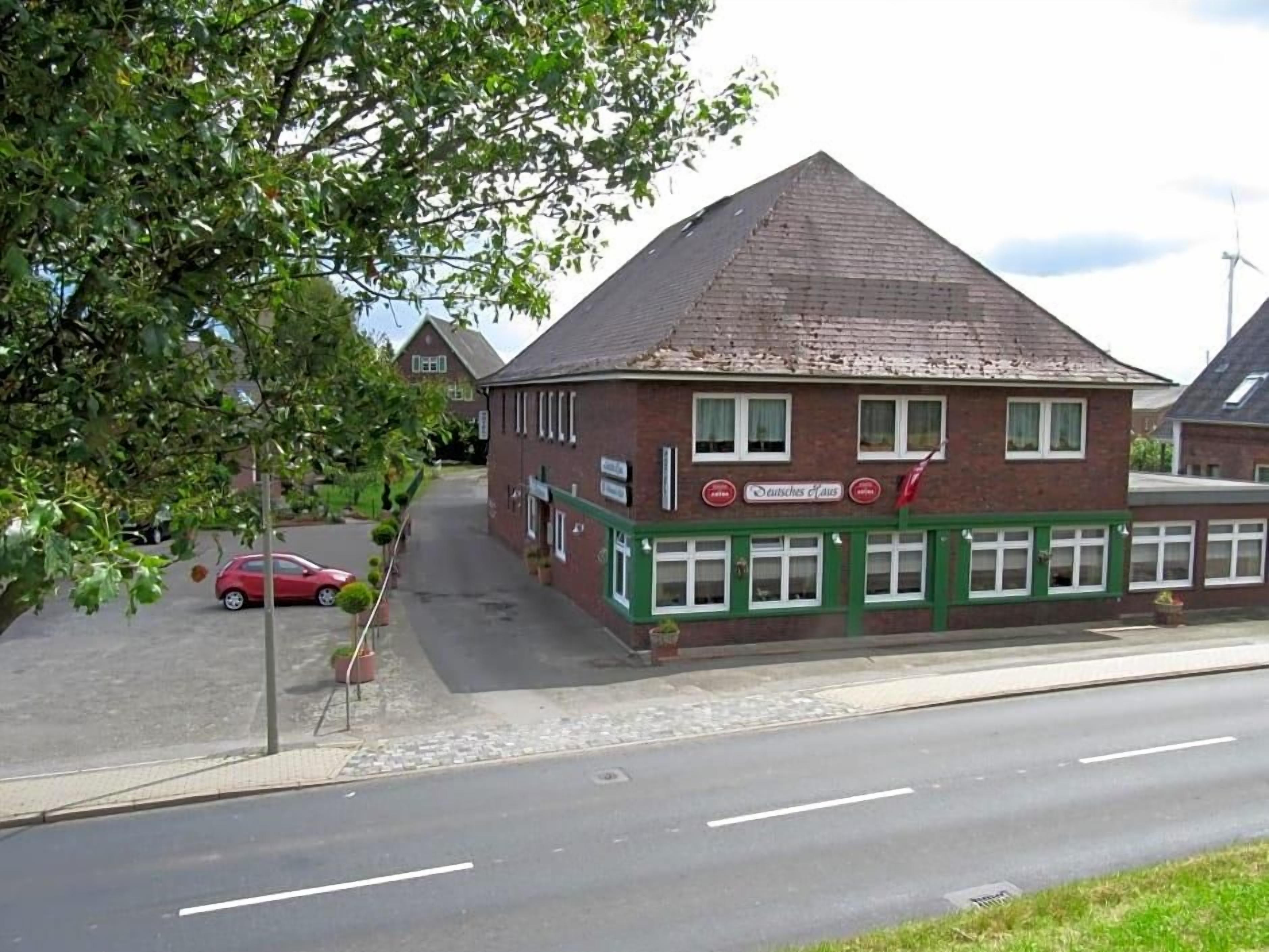 Un bâtiment à deux étages, doté d’une façade en briques et de bordures vertes, sur lequel on peut lire l’inscription « Dachtes & Haus ».