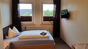 Un dormitorio con cama, dos ventanas, un radiador y un televisor en la pared.