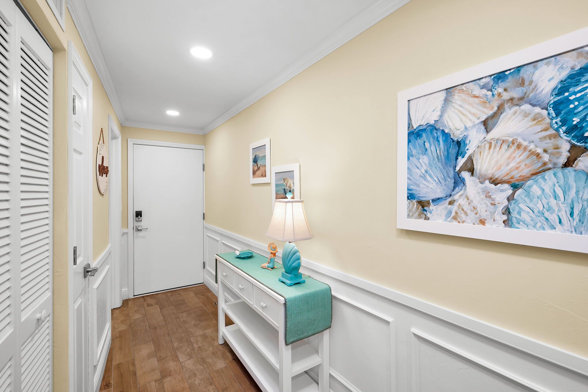 A hallway with a white door, a teal bench with a lamp, and a large seashell painting on the wall.