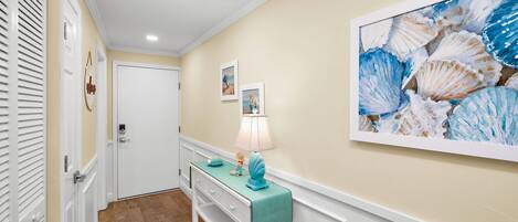 A hallway with a white door, a teal bench with a lamp, and a large seashell painting on the wall.