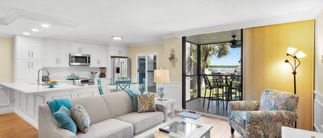 A modern living room with a sofa, coffee table, and chairs, a kitchen with white cabinets, and a view of the outdoors through a large window.