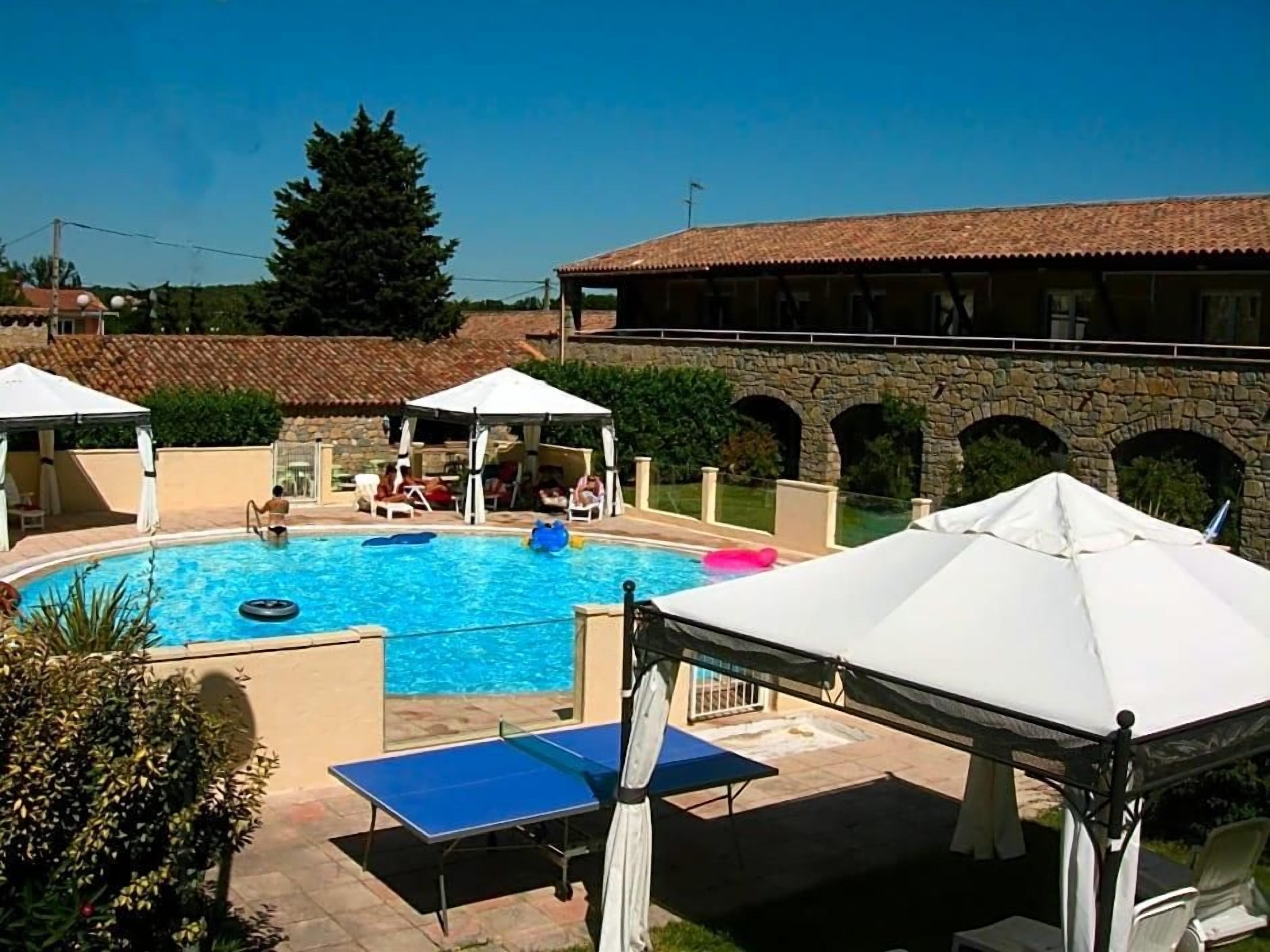 A swimming pool with lounge chairs and umbrellas, a stone bridge, and a building with a tiled roof.