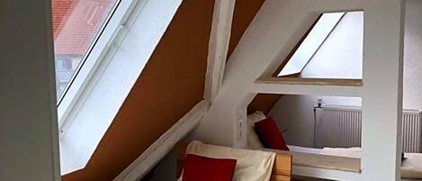 A small, compact room with sloped ceilings and skylights, featuring a single bed and a small table.
