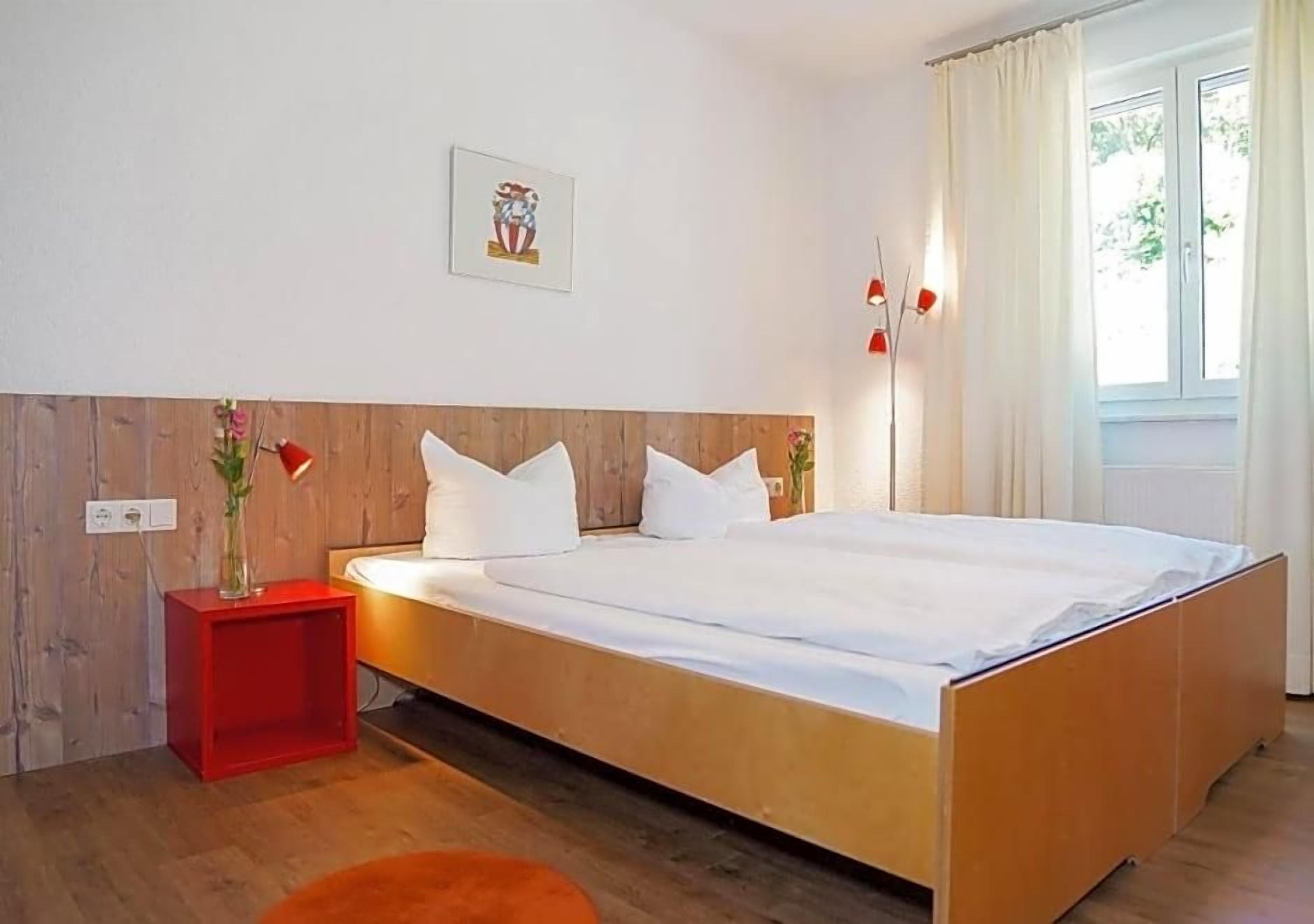 Ein modernes Schlafzimmer mit einem hölzernen Kopfende, einem Einzelbett, einem Nachttisch in Rot und einem Fenster mit Vorhängen.