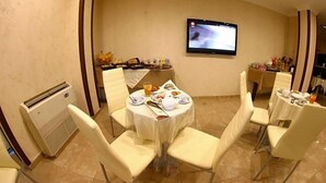 Un comedor con mesas redondas, sillas blancas, un televisor de pantalla plana en la pared y una mesa de buffet con comida y bebidas.