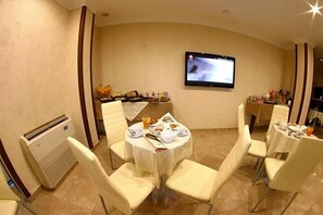 Un comedor con mesas redondas, sillas blancas, un televisor de pantalla plana en la pared y una mesa de buffet con comida y bebidas.