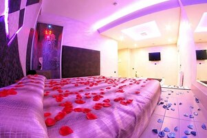 Habitación de hotel con cama cubierta de pétalos de rosa, un espejo grande e iluminación empotrada en el techo.