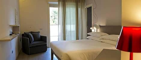 A hotel room with a bed, a desk, a chair, a bedside table, a lamp, and a window with curtains.