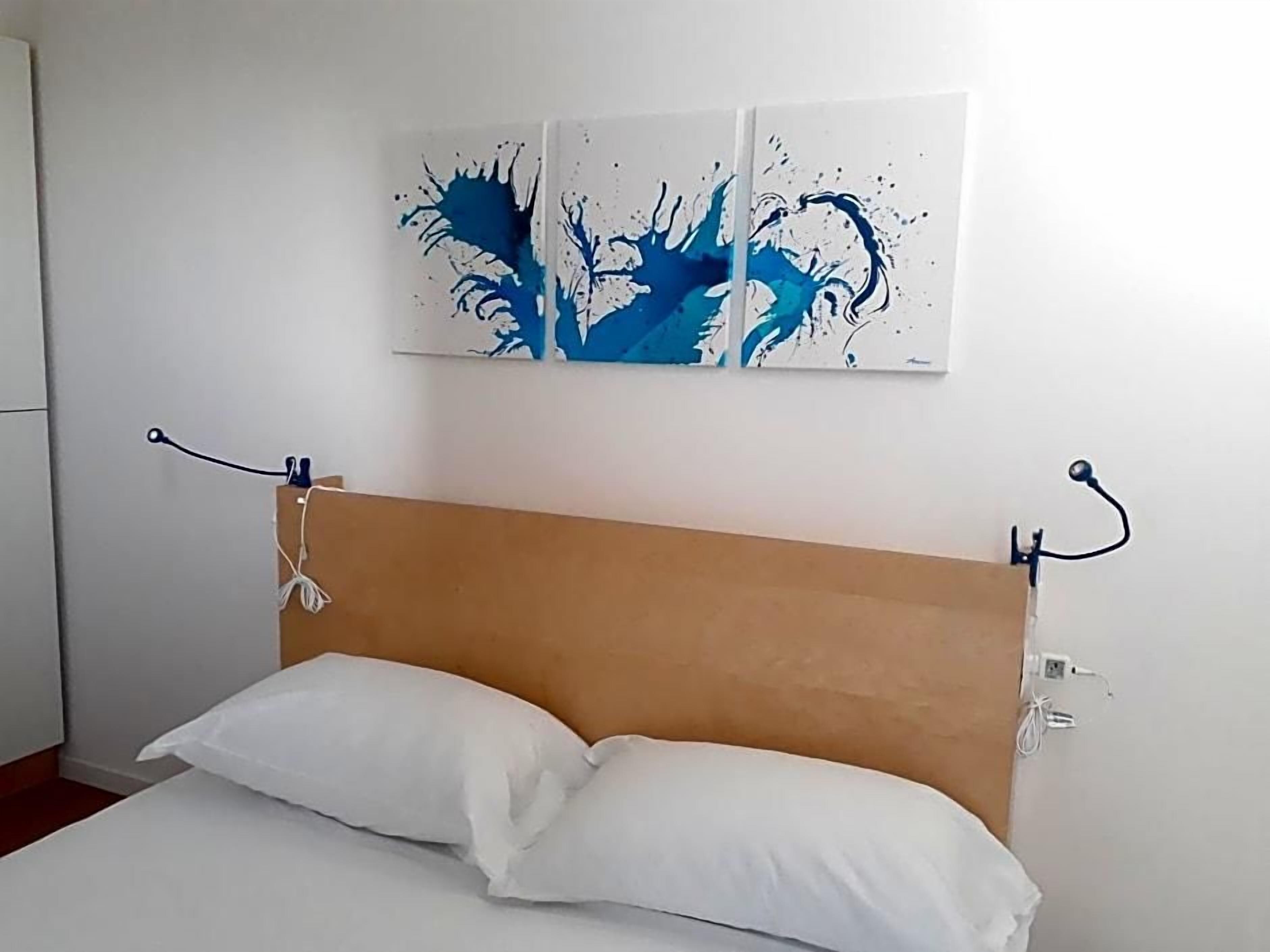 Een bed met een houten hoofdbord, wit beddengoed en een aan de muur hangend abstract schilderij.