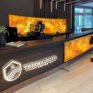 Vastaanottotiski, jossa on marmorimaisen näköinen, taustavalaistu paneeli, kahvinkeitin ja tietokonenäyttö. Hotellin nimi »THERMALHOTEL» on esillä tiskillä.