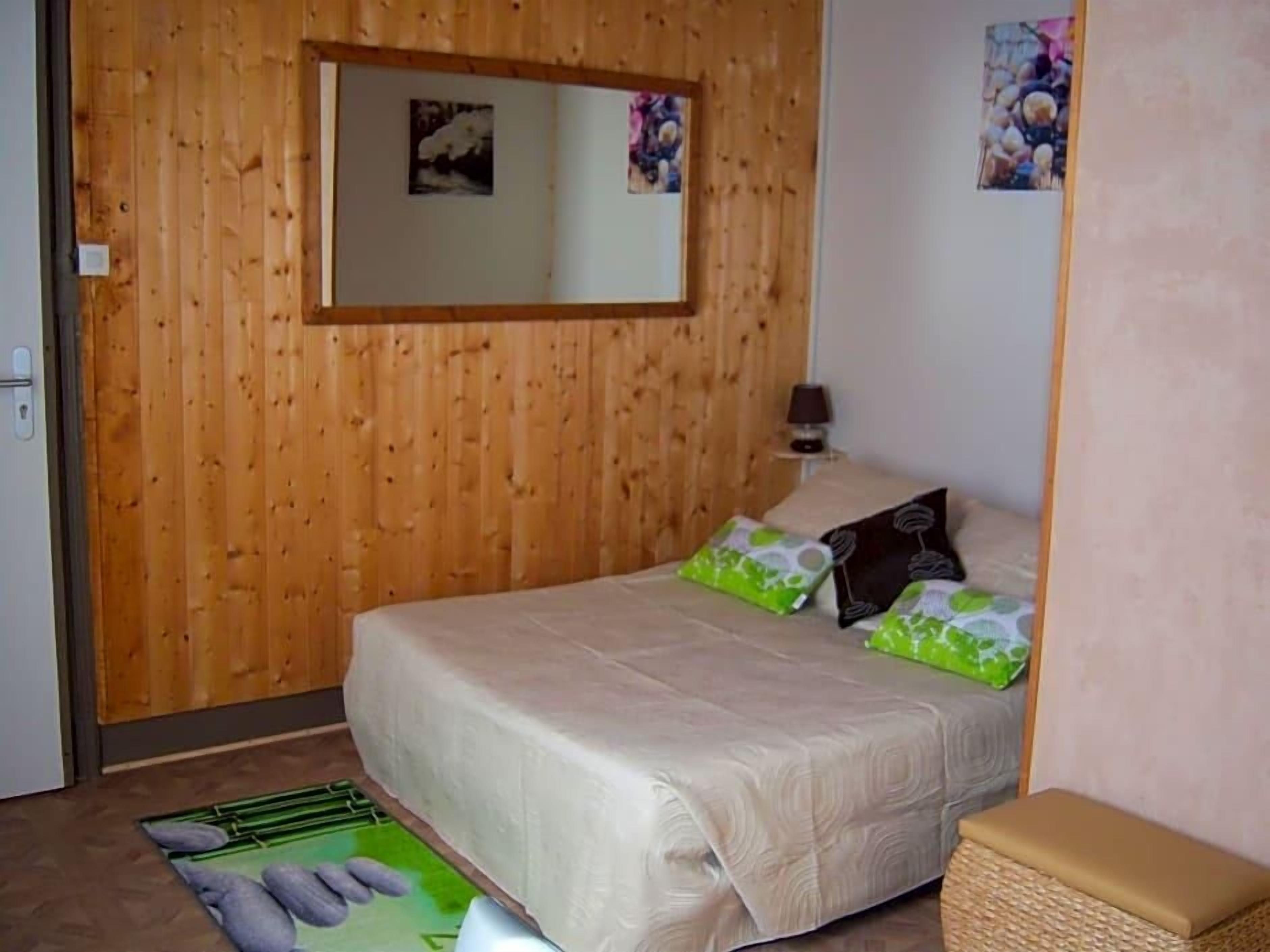 Een slaapkamer met een houten wand, een bed met een beige dekbedovertrek, een nachtkastje met een lamp en een klein spiegelbeeld aan de wand.
