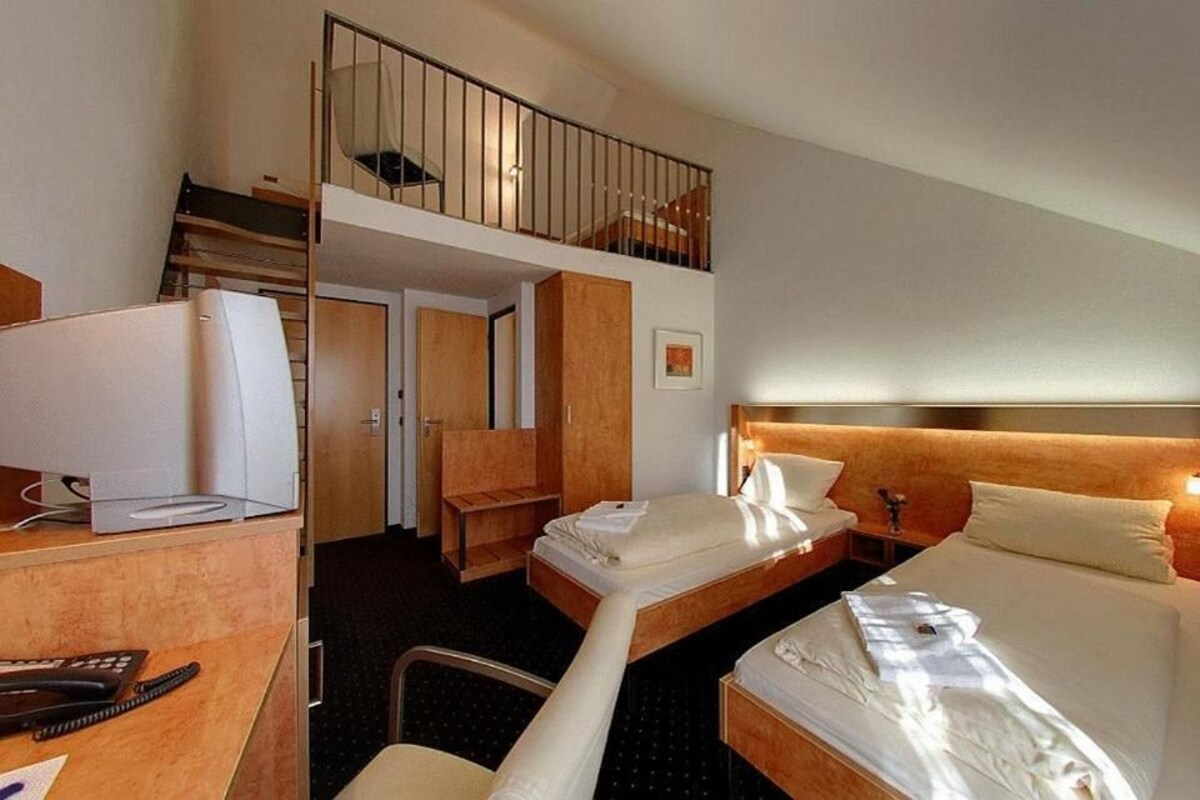 A hotel room with a desk, two beds, a chair, a wardrobe, and a staircase to an upper level.
