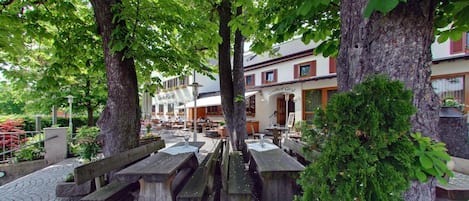 Un área de estar al aire libre con bancos y mesas de madera, rodeada de árboles y un edificio con un letrero que dice 'Gasthof Grob'.