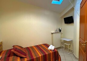 Una habitación pequeña con cama individual, colcha a rayas, una mesita con una silla, un televisor montado en la pared y un radiador.