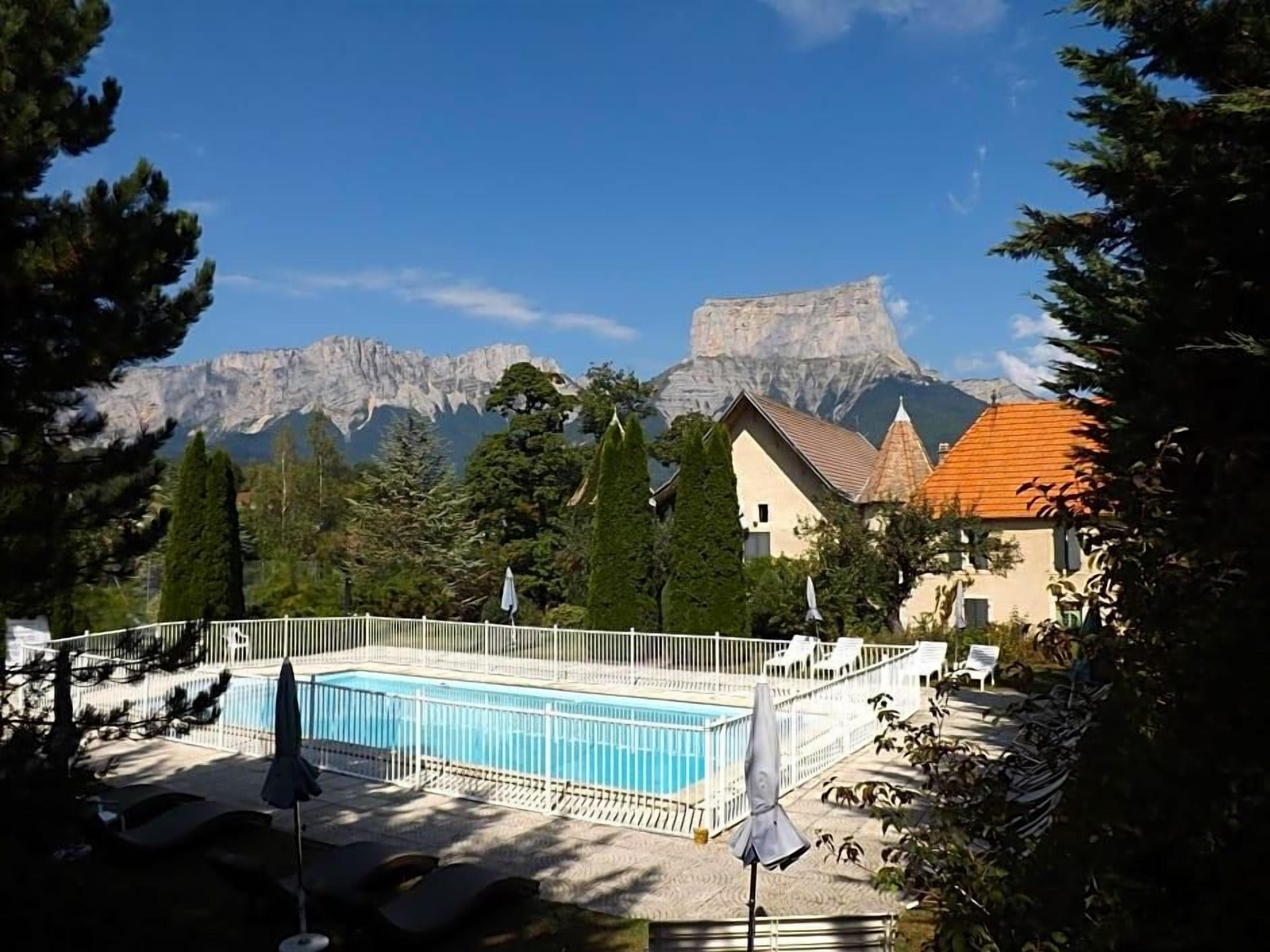 A swimming pool with a white fence, lounge chairs, and umbrellas, surrounded by trees and a mountain backdrop.