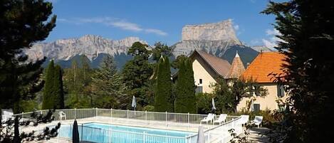 A swimming pool with a white fence, lounge chairs, and umbrellas, surrounded by trees and a mountain backdrop.