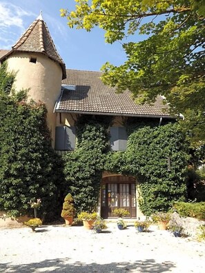 Casa de dos pisos con una torre, rodeada de follaje y plantas en macetas.
