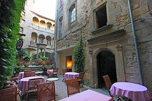 Une cour extérieure avec des tables et des chaises, des bâtiments en pierre et une fontaine.