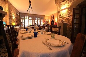 A dining room with a round table set for a meal, stone walls, and a view through large windows. - H Otel Restaurant D Alibert (Caunes-Minervois)
