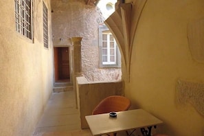 Un pasillo estrecho con una mesa y una silla, una ventana con barrotes y una puerta que da a otra habitación.