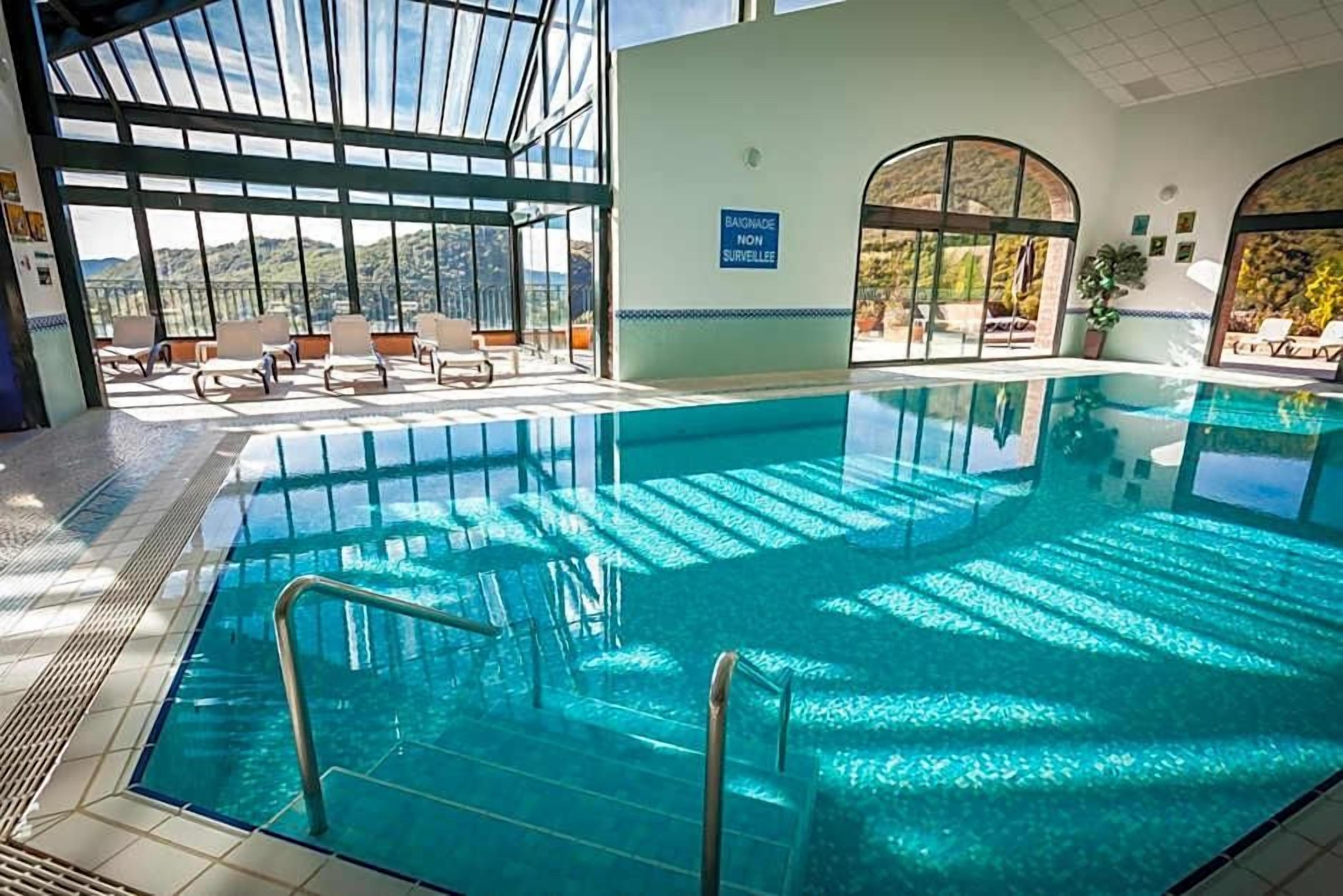 An indoor swimming pool with clear glass walls, a sunlit area with lounge chairs, and a sign indicating the pool is not monitored.