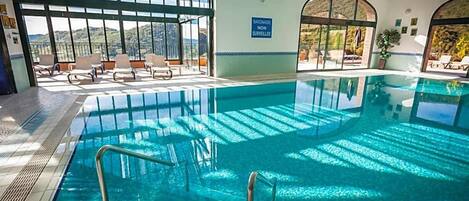 An indoor swimming pool with clear glass walls, a sunlit area with lounge chairs, and a sign indicating the pool is not monitored.