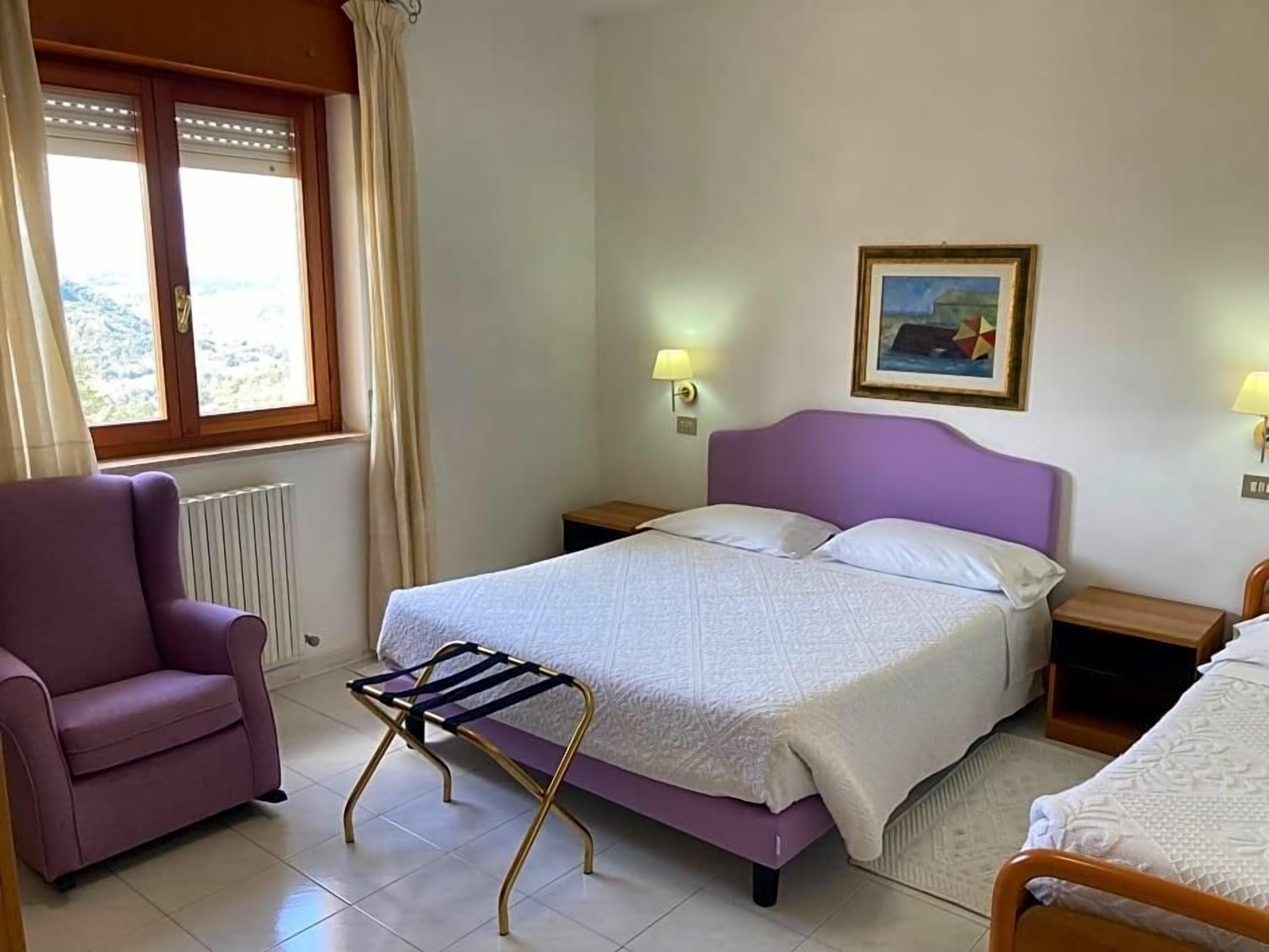 A bedroom with a bed, a chair, a radiator, a window with curtains, and a painting on the wall.