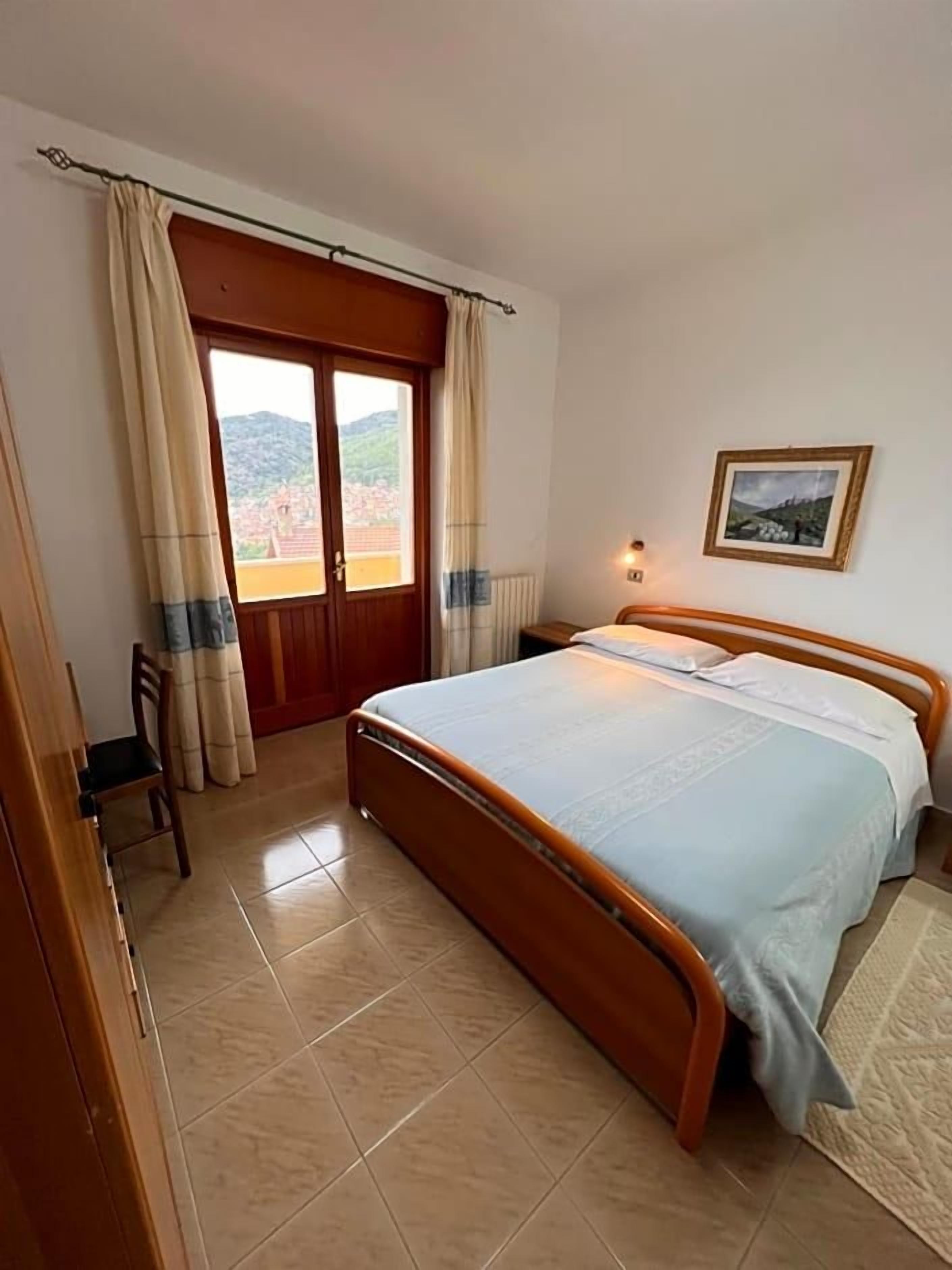 Una camera da letto con un letto, tende, una sedia, un termosifone e un quadro appeso al muro.